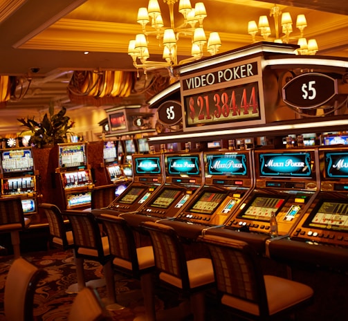 A casino floor featuring rows of brightly lit video poker machines. The area is illuminated by elegant chandeliers, with a display board showing a large sum of money, suggesting a possible jackpot. The rich decor includes luxurious carpets and comfortable seating, adding to the opulent atmosphere.
