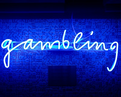 A bright blue neon sign spells out the word 'gambling' in cursive script. The background features a textured wall covered in domino tiles, also illuminated by the blue light, creating a vivid and visually striking scene.