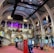 The interior of a large, ornately designed building with arched ceilings and intricate architectural details. The space features gaming machines lined up along the floor, under a high ceiling with a grid pattern. Lighting in purple and white tones enhances the ambiance.