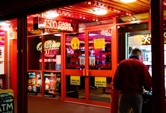 A brightly lit entrance to a casino with neon signs, including one advertising a £500 jackpot. The doors have clear signage indicating entry is restricted to those over 18. A person in a red jacket stands to the right, possibly interacting with a machine or display.