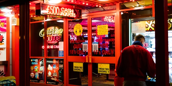 A brightly lit entrance to a casino with neon signs, including one advertising a £500 jackpot. The doors have clear signage indicating entry is restricted to those over 18. A person in a red jacket stands to the right, possibly interacting with a machine or display.