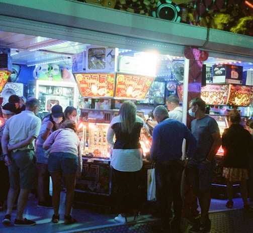 A group of people standing in front of an illuminated amusement arcade, engaging with various gaming machines. The environment is bustling with activity, and colorful lights create a vibrant atmosphere. The ceiling is decorated with large, playful images of dice and other game-related graphics, enhancing the lively mood.