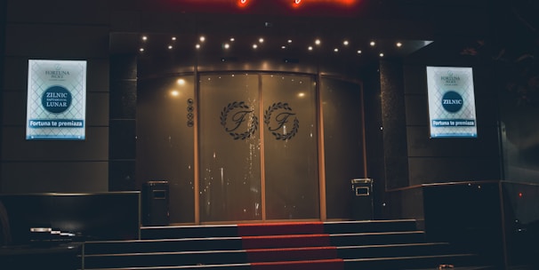 A neon-lit casino entrance with a red carpet leading up to large double doors. The sign reads 'Fortuna Palace Luxury Games' in bright red and white neon lights. The dimly lit atmosphere suggests a night-time setting. There are two digital displays on either side of the entrance showing promotional content.