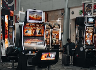 A collection of slot machines is situated inside a building with bright, colorful screens displaying various games and gaming themes. A person is seated nearby, seemingly relaxed with luggage beside them, and the atmosphere is lively with electronic displays.