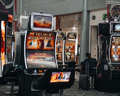 A collection of slot machines is situated inside a building with bright, colorful screens displaying various games and gaming themes. A person is seated nearby, seemingly relaxed with luggage beside them, and the atmosphere is lively with electronic displays.